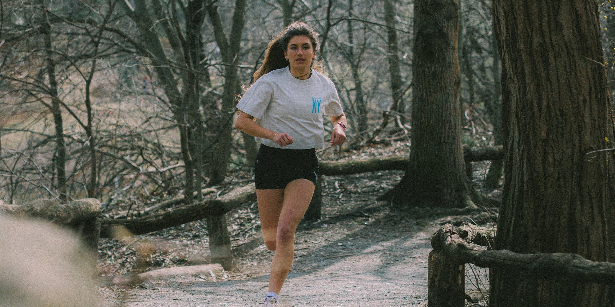 Female Athlete wearing a cropped off-top shirt with "NY" logo in blue, running on a trail.