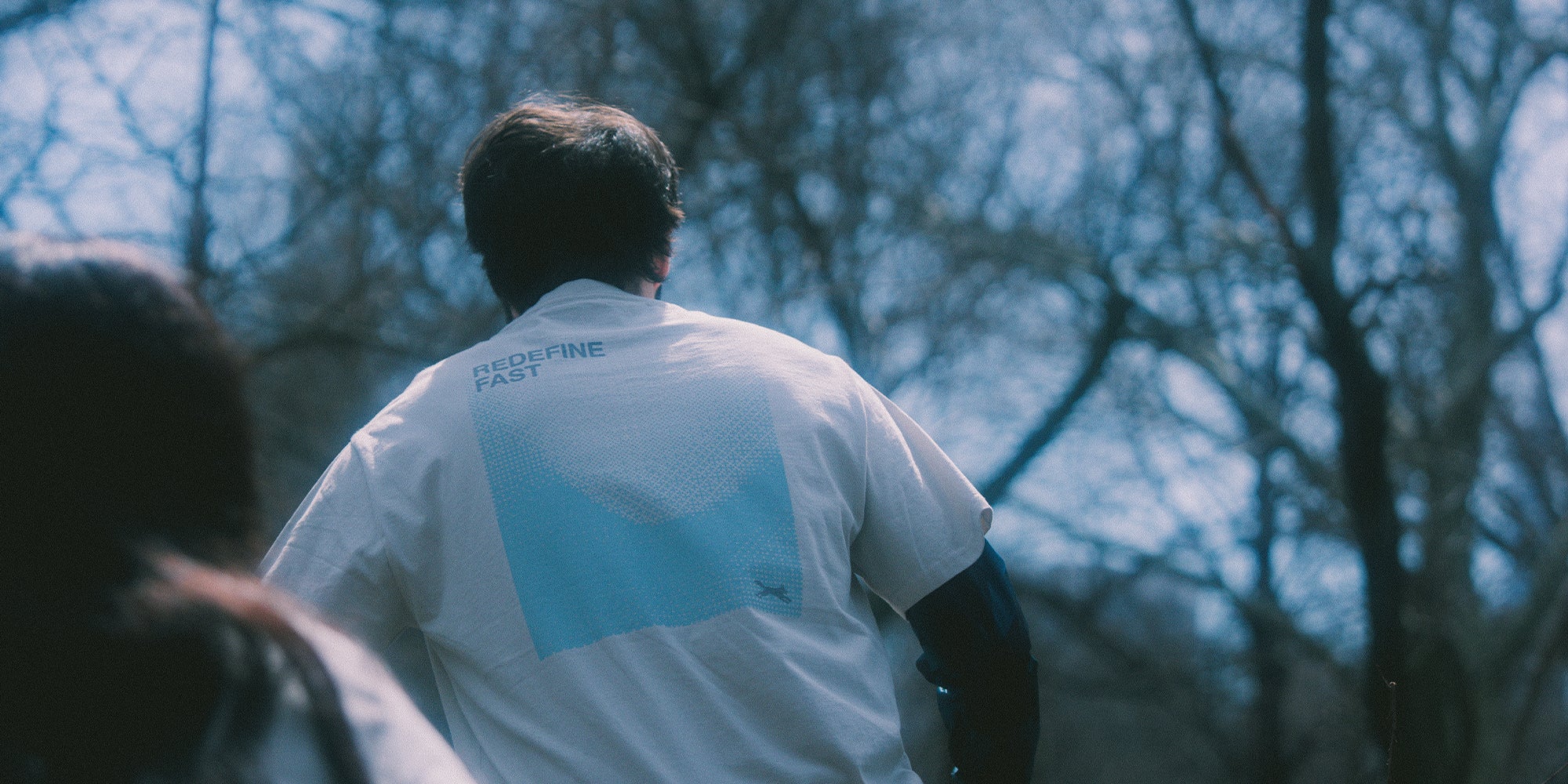 Person running and wearing a white shirt with a blue logo Redefine fast artistic print in a forest setting.
