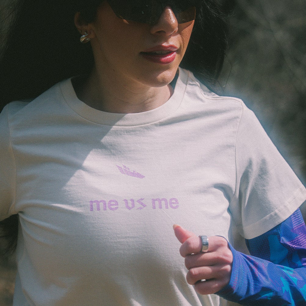 Female Athlete wearing an off-white shirt with "me vs me" logo on the front in purple gothic font.